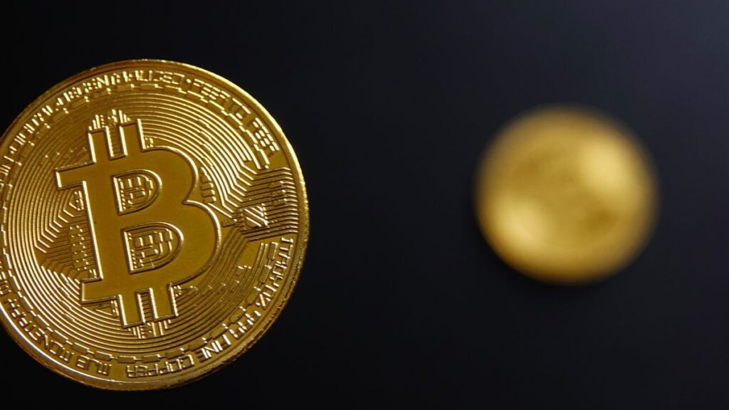 A gold coin with a &quot;B&quot; symbol is shown in focus on the left-hand side of the image. The background is black and blurred. On the right-hand side of the image there is another, smaller blurred gold coin.